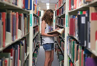 woman standing in the book stacks looking at an open book